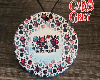 May include: A round, white ornament with a black string. The ornament features a colorful illustration of various cryptids, gifts, and snowflakes, with the text "merry cryptid". The ornament has the text "CARO GREY" in red.
