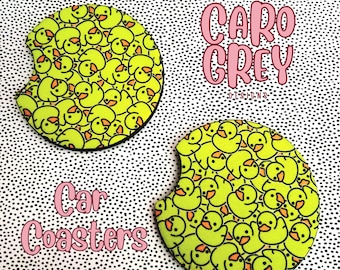 May include: Two yellow rubber ducky car coasters with a black outline. The coasters are on a white background with black polka dots. The text "Caro Grey Designs" is in pink letters on the top right of the image. The text "Car Coasters" is in pink letters in the bottom left of the image.
