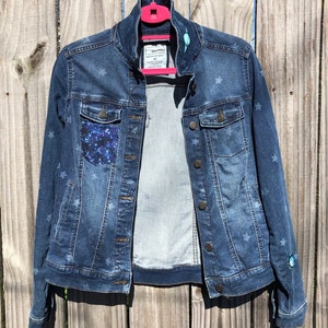 Puede incluir: Una chaqueta vaquera azul con estrellas blancas impresas por todas partes. La chaqueta tiene un bolsillo azul brillante en el lado izquierdo y un pequeño parche de estrella azul en la manga derecha. La chaqueta está colgada en una percha rosa.