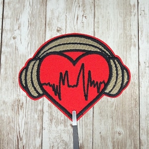 May include: A red heart-shaped patch with a black outline and a black heartbeat line. The heart is framed by tan and black headphones. The patch is likely for clothing or accessories.