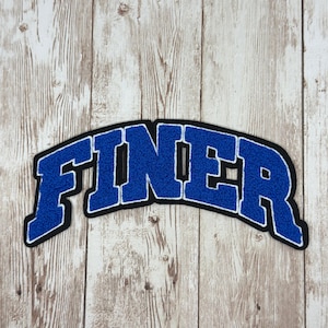 May include: A blue felt patch with the word "FINER" in a bold, arched font. The letters are outlined in black and have a white border. The patch is set against a light wood grain background.