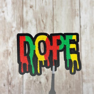May include: Embroidered patch with the word "DOPE" in red, yellow, and green, outlined in black. The letters have a dripping effect. The patch is on a light wood grain background.