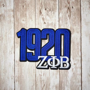 May include: An embroidered patch featuring the blue numbers 1920 with a black border, positioned above the white Greek letters Zeta Phi Beta, which are also outlined in black, creating a bold and clear design for fabric application.