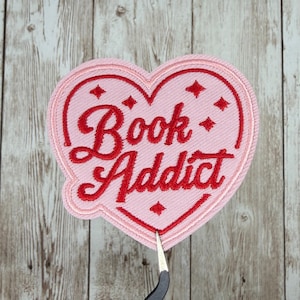 May include: A pink heart-shaped patch with the words "Book Addict" embroidered in red script. The patch has a red border and four red diamond accents. The background is a light pink color.