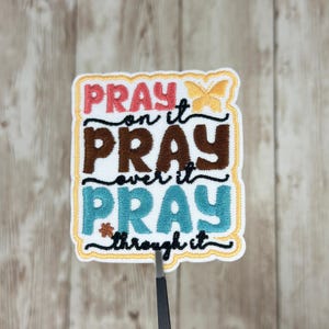 May include: Embroidered patch with the words "PRAY on it, PRAY over it, PRAY through it" in pink, brown, and teal. A yellow butterfly and flower accents the design. The patch has a white background with a yellow border.