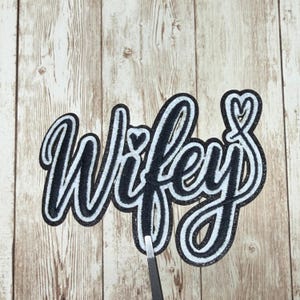 May include: A black and white patch with the word "Wifey" in a cursive font. The patch has a black outline and a white interior, with small heart details. The patch is held by tweezers against a wooden background.