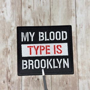 Pode incluir: Patch quadrado preto com texto branco que diz "MY BLOOD TYPE IS BROOKLYN". As palavras "TYPE IS" são vermelhas. O patch está sobre uma superfície de grão de madeira clara.