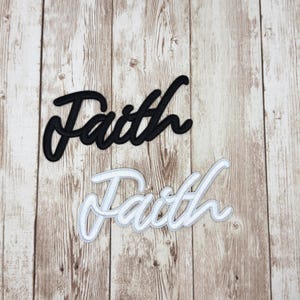 May include: Two embroidered patches with the word "Faith" in a cursive script. One patch is black with a black border, and the other is white with a white border. The patches are on a light brown wooden surface.