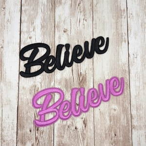 May include: Two embroidered patches with the word "Believe" in a cursive font. One patch is black, and the other is a vibrant pink. The patches are displayed on a light brown wooden surface.