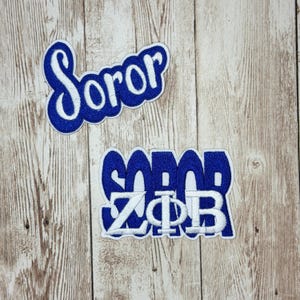 以下が含まれることがあります： 青い筆記体でSororと刺繍された2つのワッペン。白い縁取りが施されています。下のワッペンには、白い背景の青い文字の上に重ねられた白いブロック体のギリシャ文字Zeta Phi Betaも含まれています。
