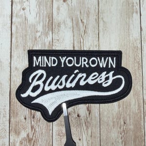May include: Black embroidered patch with the words "MIND YOUR OWN Business" in white script. The patch has a black border and is rectangular with rounded edges. The patch is on a light brown wooden surface.