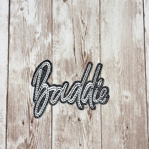 May include: A black and white embroidered patch with the word "Baddie" in a cursive font. The patch has a glittery black background with white stitching around the edges. The patch is set against a light brown wood grain background.
