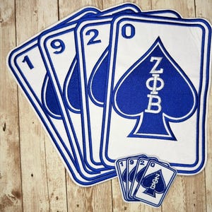 May include: A set of four white and blue embroidered patches shaped like playing cards, each featuring a number from the sequence 1, 9, 2, 0. The final card displays a blue spade symbol with the white Greek letters Zeta Phi Beta centred inside.