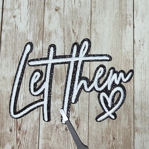 May include: Black and white embroidered patch with the words "Let Them" and a heart symbol. The patch has a glittery black border and white stitching. The patch is on a light brown wooden surface.
