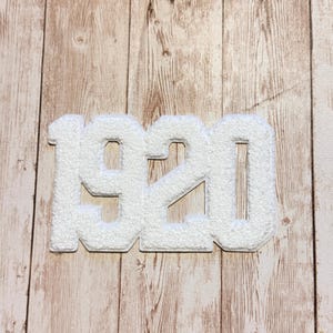 Puede incluir: Un parche de chenilla blanca con los números 1920 en una fuente de bloque en negrita. El parche tiene una superficie texturizada y difusa con un borde troquelado limpio, diseñado para aplicarse sobre superficies de tela.