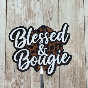 May include: Embroidered patch with the words "Blessed & Bougie" in white script with a black outline. The background is a brown and black leopard print. The patch is held by tweezers.