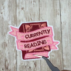 Puede incluir: Parche bordado con un libro burdeos y una pancarta rosa que dice "CURRENTLY READING". La pancarta tiene pequeños detalles de corazones. El parche tiene un borde blanco.