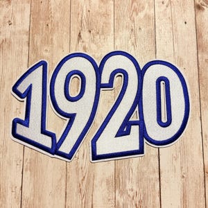 Könnte beinhalten: Ein weißer Stickaufnäher mit der Zahl 1920 in einer fetten Blockschrift. Die Ziffern sind mit einem dicken, dunkelblauen Rand versehen, was für einen kontrastreichen Look sorgt, der sich für die Applikation auf verschiedenen Stoffen eignet.