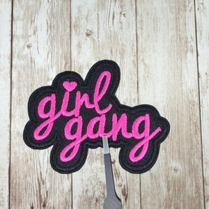 May include: Embroidered patch with the words "girl gang" in pink script lettering on a black background. A small pink heart is above the word "girl". The patch is held by a pair of tweezers against a wooden background.