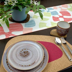 May include: A cork placemat with a red felt insert and two ceramic plates. The plates have a white glaze with brown speckles. Two silver forks and a silver spoon are on the placemat.