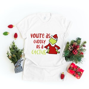 May include: White t-shirt with the text "you're as cuddly as a cactus" and a cartoon Grinch character wearing a Santa hat and red suit. Christmas-themed decor surrounds the shirt.