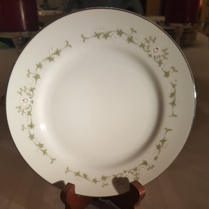 May include: A white ceramic dinner plate with a green and white floral design. The plate has a silver rim and is on a wooden stand.