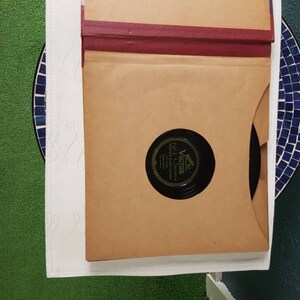 May include: A brown cardboard record sleeve with a red spine. The sleeve is open and a black vinyl record is visible inside. The record label is black with white text that reads "Victor" and "The Victor Talking Machine Company".