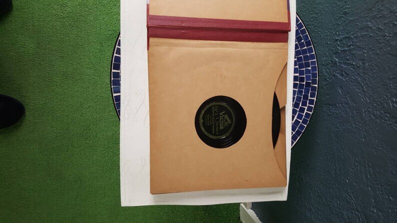 May include: A brown cardboard record sleeve with a red spine. The sleeve is open and a black vinyl record is visible inside. The record label is black with white text that reads "Victor" and "The Victor Talking Machine Company".