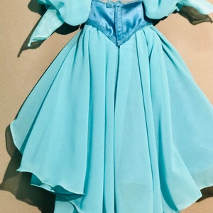 Op de afbeelding: Een lichtblauwe jurk met een gelaagde rok en pofmouwen. Het lijfje is een donkerdere tint blauw met een satijnen afwerking en kanten bies. De jurk is voor een kind.