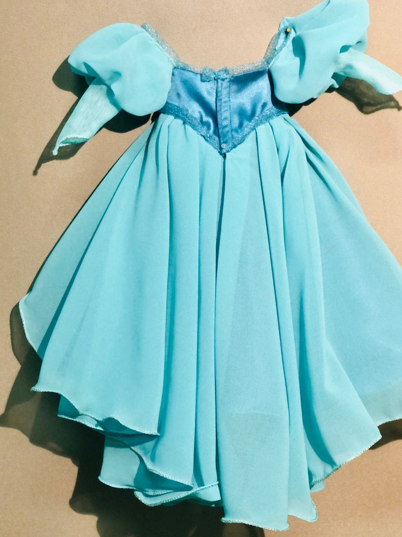 Op de afbeelding: Een lichtblauwe jurk met een gelaagde rok en pofmouwen. Het lijfje is een donkerdere tint blauw met een satijnen afwerking en kanten bies. De jurk is voor een kind.