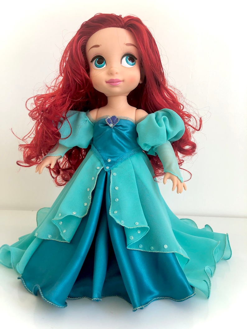 Op de afbeelding: Pop van een personage met rood haar en blauwe ogen, gekleed in een teal en turquoise jurk. De jurk van de pop heeft een gelaagd ontwerp met een satijnen rok en een doorschijnende overlay, versierd met kleine, sprankelende versieringen. Het haar van de pop is gestyled in losse golven.