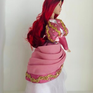 May include: A porcelain doll with long red hair wearing a pink and white dress with gold trim. The dress has a ruffled skirt and a fitted bodice.