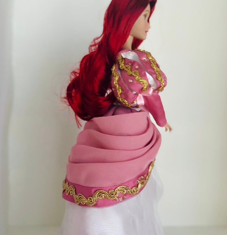 May include: A porcelain doll with long red hair wearing a pink and white dress with gold trim. The dress has a ruffled skirt and a fitted bodice.