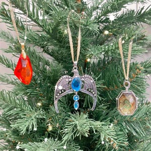 May include: Three Christmas ornaments hanging on a green artificial Christmas tree. The ornaments are silver with blue and red gems. One ornament is a crown with wings, one is a red gem, and one is a locket with a silver snake inside.