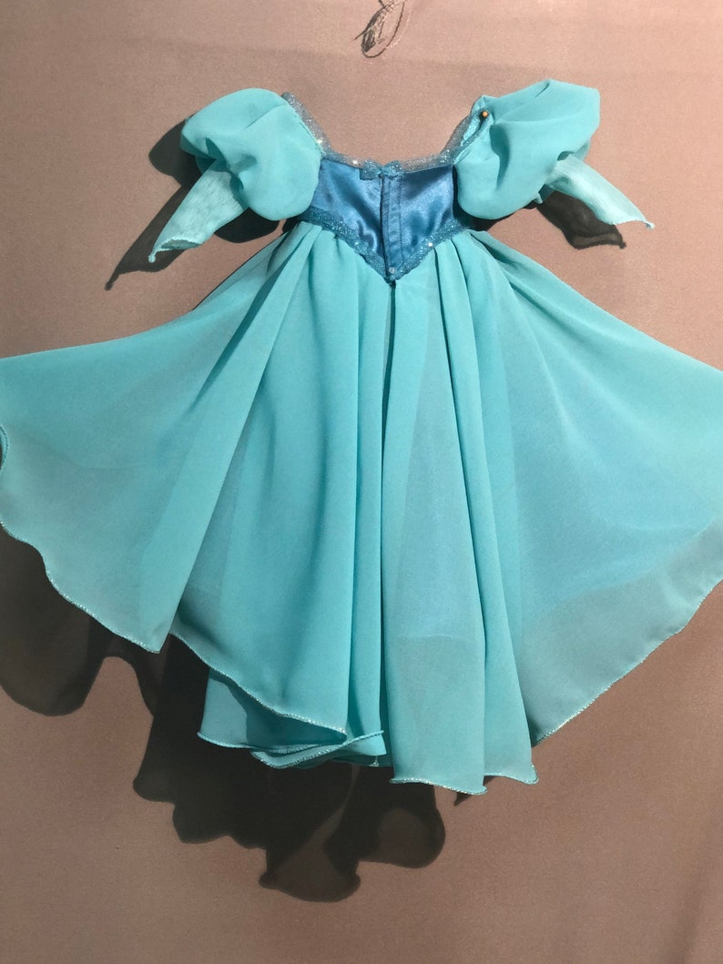 Op de afbeelding: Een lichtblauwe jurk met een zwierige rok en pofmouwen. Het lijfje is een donkerdere tint blauw met pailletten. De jurk lijkt gemaakt van een lichte, transparante stof, geschikt voor een kostuum of speciale gelegenheid.