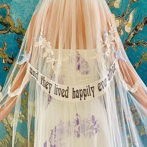 May include: A white tulle wedding veil with a white satin ribbon that says "and they lived happily ever after". The veil is decorated with blue and white floral embroidery.