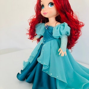 Op de afbeelding: Pop van een personage met felrood haar en grote blauwe ogen. De pop draagt een lichtblauwe en blauwgroene jurk met een gelaagde rok en off-the-shoulder mouwen. Een klein juweel siert de halslijn.