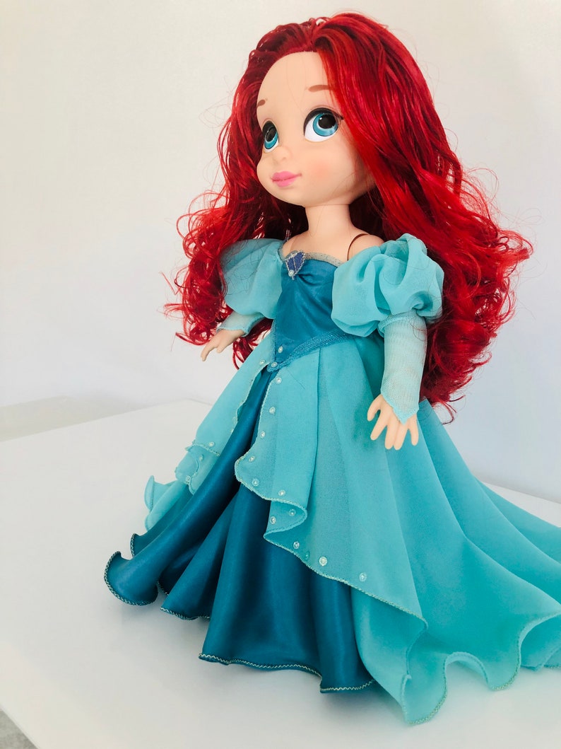 Op de afbeelding: Pop van een personage met felrood haar en grote blauwe ogen. De pop draagt een lichtblauwe en blauwgroene jurk met een gelaagde rok en off-the-shoulder mouwen. Een klein juweel siert de halslijn.