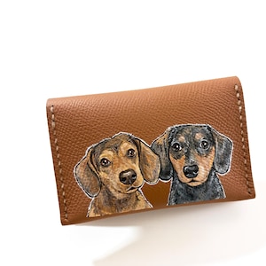 May include: A brown leather wallet with a hand-painted image of two dachshunds. One is brown and tan, the other is black and brown. The wallet has white stitching around the edges.