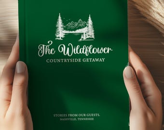 May include: A green journal titled "The Wildflower Countryside Getaway" held by a person wearing a white sweater. The cover features a white illustration of trees, mountains, and the text "Stories from our guests. Nashville, Tennessee."