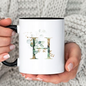 May include: White ceramic mug with a black handle and a gold and green floral design featuring the letter "H".