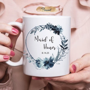 Może przedstawiać: Biały kubek ceramiczny z wzorem niebieskiego wieńca kwiatowego w akwareli. Tekst "Maid of Honor 8.14.25" jest nadrukowany czarnym kolorem wewnątrz wieńca.