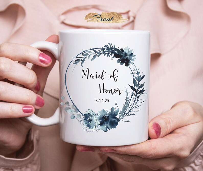 Może przedstawiać: Biały kubek ceramiczny z wzorem niebieskiego wieńca kwiatowego w akwareli. Tekst "Maid of Honor 8.14.25" jest nadrukowany czarnym kolorem wewnątrz wieńca.