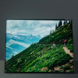 May include: A framed photograph of a mountain landscape with a person hiking on a trail. The photo features a green hillside with white flowers, a winding path, and a distant mountain range with snow-capped peaks.