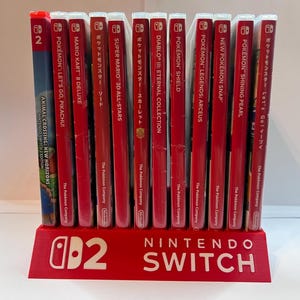 May include: A red plastic Nintendo Switch game case stand holding twelve video game cases in a row. The stand features the white Nintendo Switch logo and the number 2. The game titles include Animal Crossing, Mario Kart, and various Pokémon games.