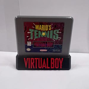 Könnte beinhalten: Eine graue Mario's Tennis Videospielkassette für die Virtual Boy Konsole. Das Etikett zeigt den Titel in grünem und gelbem Text, einen gelben Tennisball und das Virtual Boy Logo. Die Kassette steht in einem schwarzen Ständer mit VIRTUAL BOY in Rot.