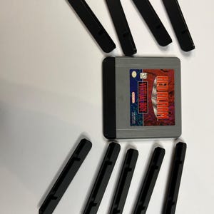 Könnte beinhalten: Ein graues Virtual Boy-Spielmodul mit dem Titel Red Alarm und einem rot-schwarzen Etikett, umgeben von acht schwarzen Staubschutzhüllen aus Kunststoff, die in zwei Reihen über und unter dem Spielmodul auf weißem Hintergrund angeordnet sind.