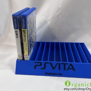 以下が含まれることがあります： 複数の垂直スロットを備えた青いプラスチック製のゲームケース用収納ラック。前面には黒い文字でPS VITAとPlayStation Vitaと印字されています。ラックの左側には、日本語のテキストとGod Eater 2のタイトルが記載されたゲームケースが2つ差し込まれています。