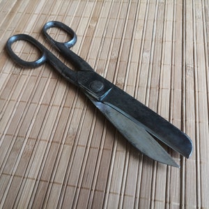 May include: A pair of vintage metal shears with a black finish. The shears have a long blade and a shorter blade. The shears are lying on a bamboo mat.