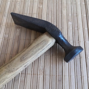 May include: A metal hammer featuring a dark, textured iron head with a flat, wide striking surface and a curved, tapered end. The head is securely attached to a light-colored, straight wooden handle with a visible grain pattern throughout its length.